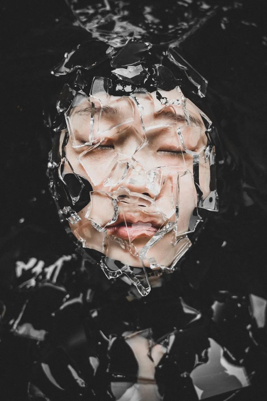 黑色液体般的背景前,一张闭着眼睛的人脸被破碎玻璃和反光碎片局部遮挡。